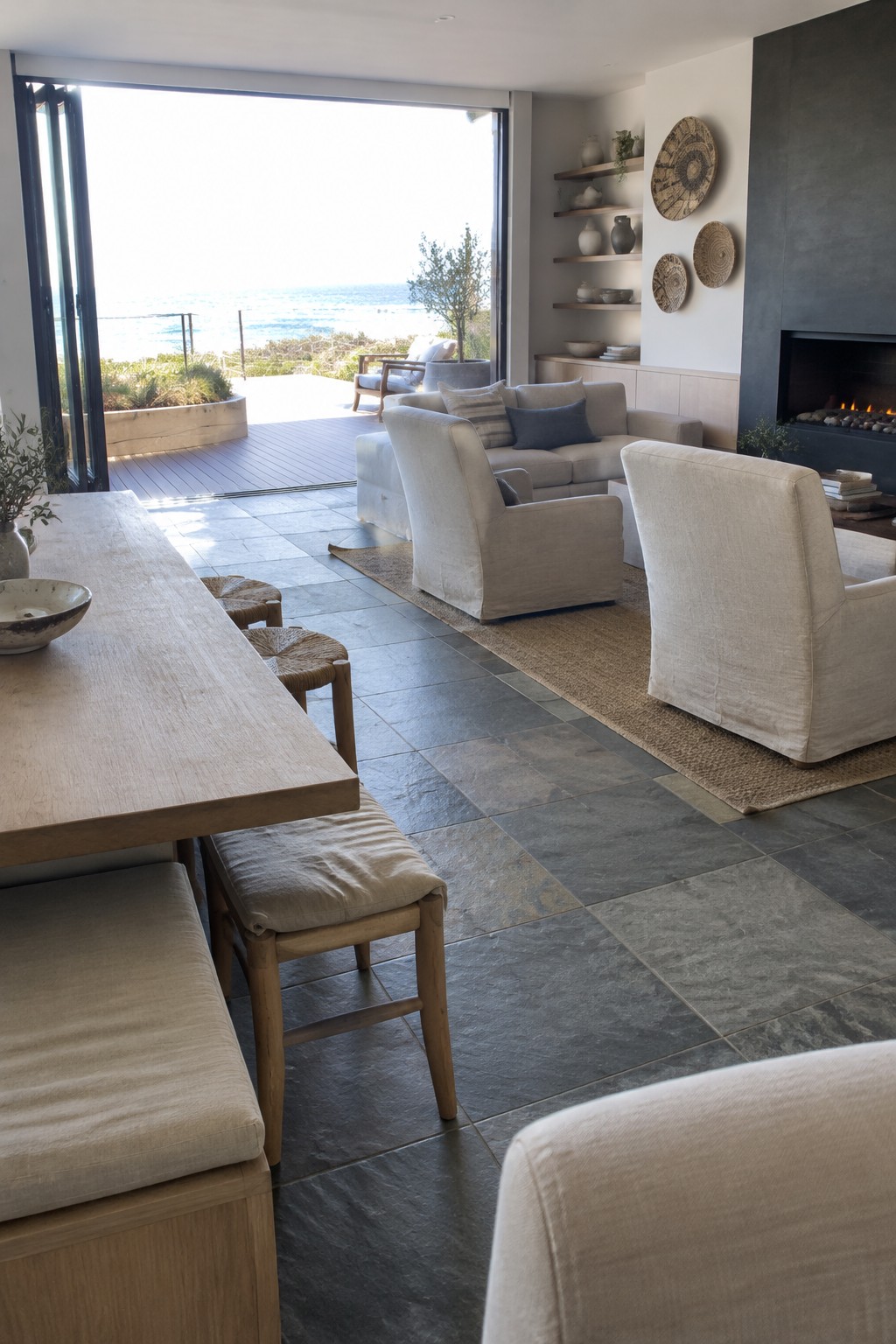 A coastal living and dining room with a sofa and armchairs on a rug beside a long wooden dining table and open glass doors.