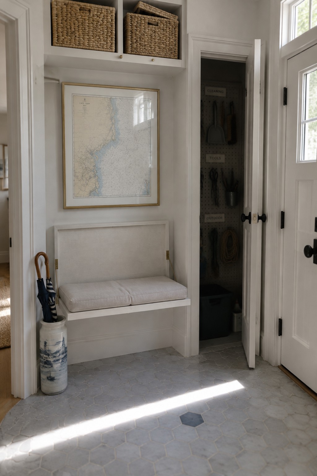 A white entryway with a built-in bench beneath a framed map, baskets on an upper shelf, and an open closet with cleaning tools.