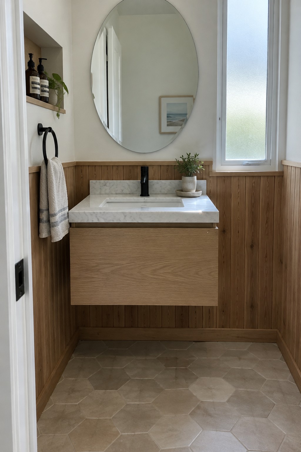 A bathroom vanity with a wood cabinet and paneled lower walls, a marble countertop, an oval mirror, and a frosted window.
