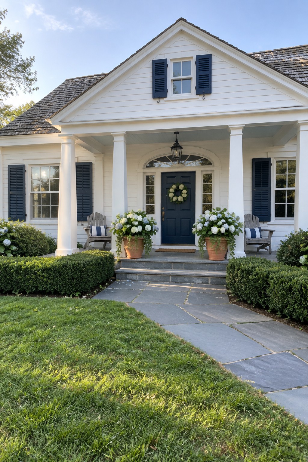 White coastal-style house with a dark blue front door, columned porch, shuttered windows, flower planters, and a stone walkway.