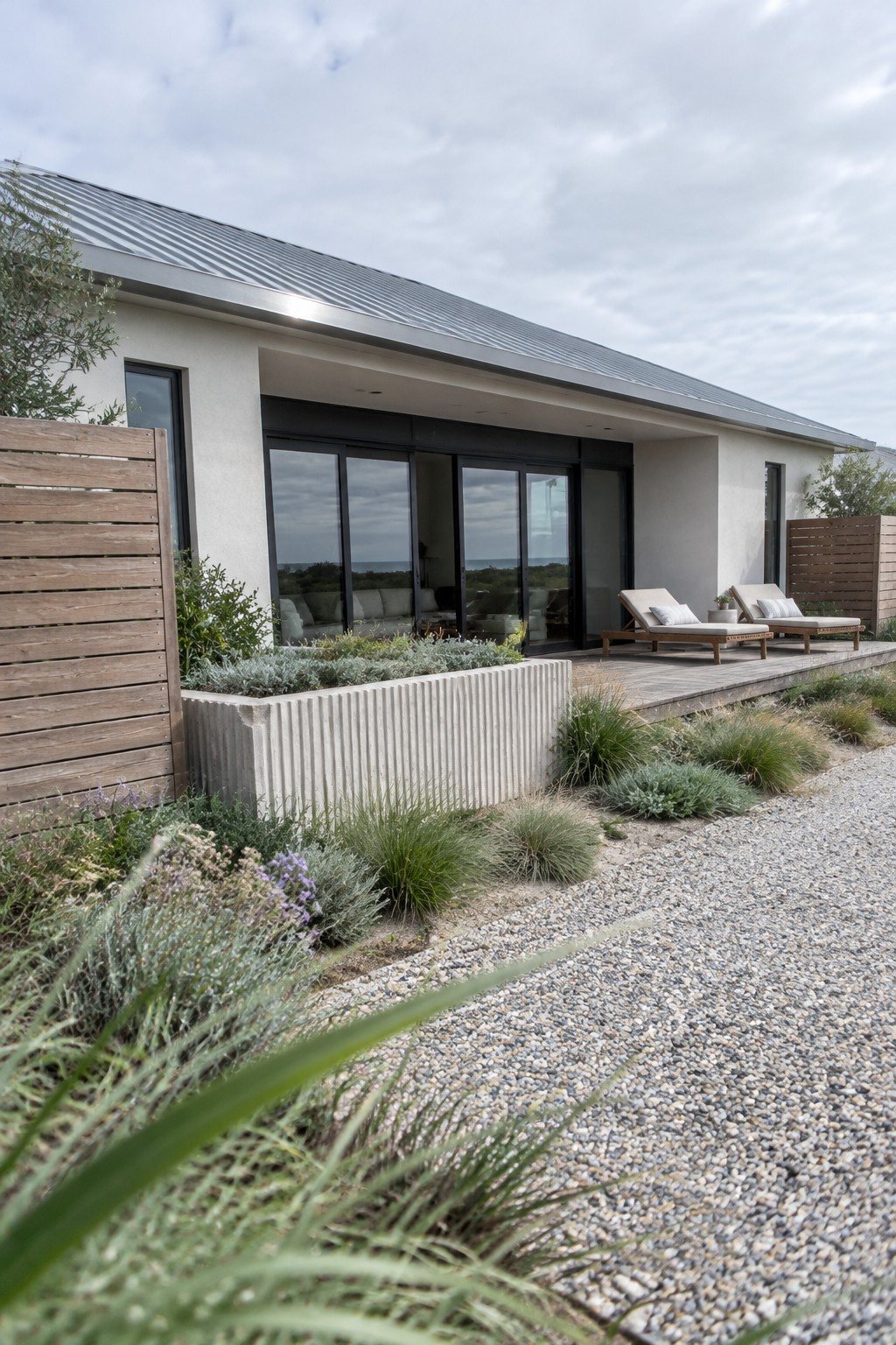 A single-story house with a deep roof overhang, sliding glass doors, a wood deck, and two outdoor lounge chairs.