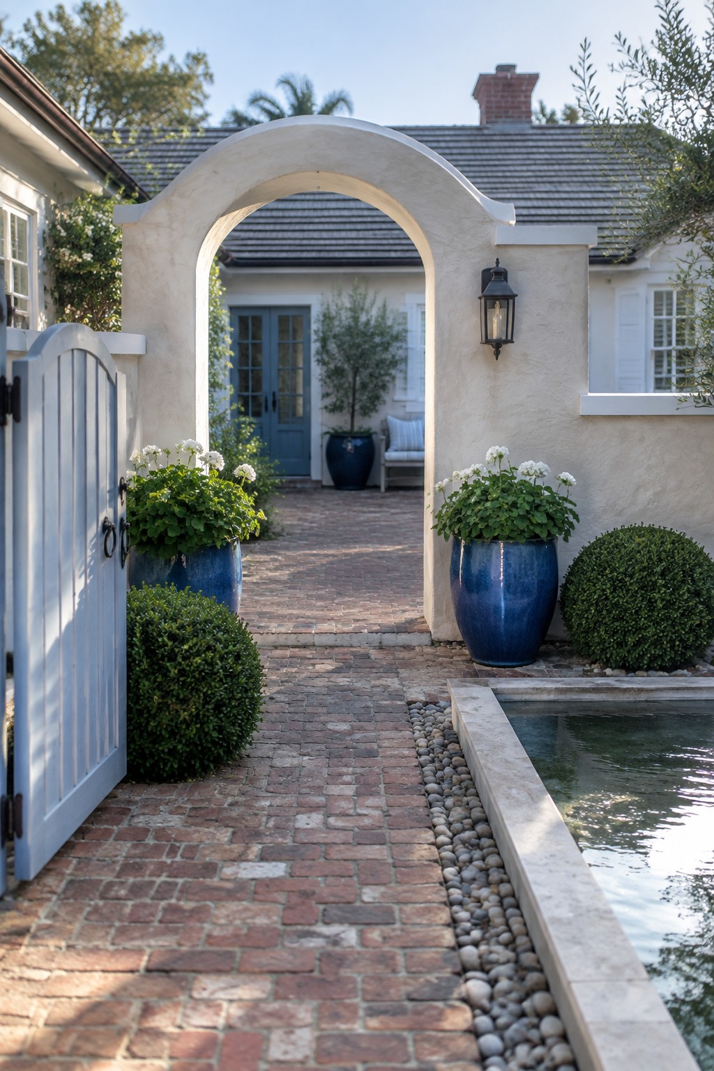 A brick walkway passes through a stucco archway toward a blue front door, with an open white gate, potted flowers, and a narrow pool beside the path.