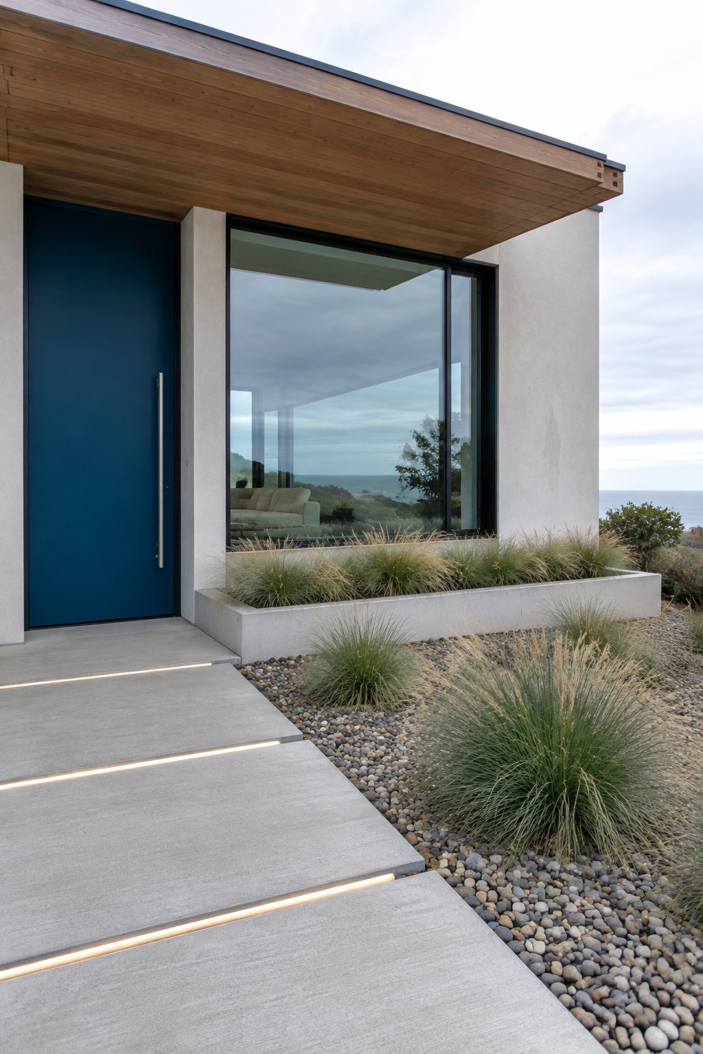 A deep blue front door and large black-framed window set into a pale stucco coastal home beneath a wood soffit.