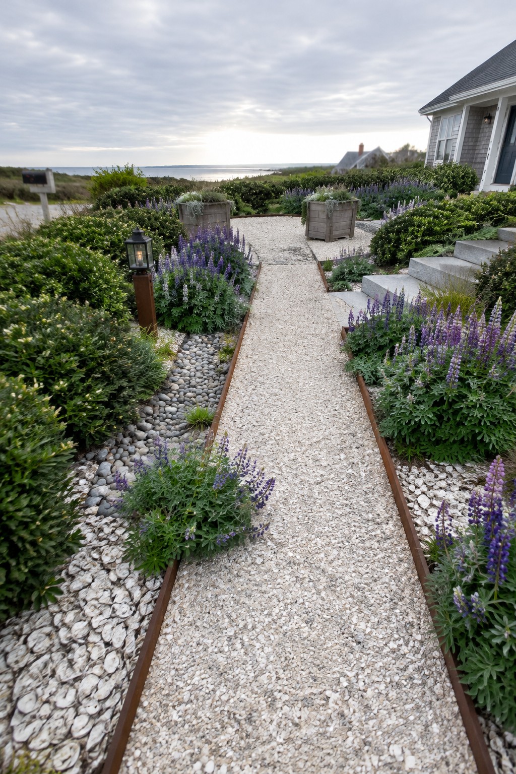 A gravel walkway with metal edging runs through coastal garden beds toward a small gravel landing, with purple flowers and shrubs on both sides.