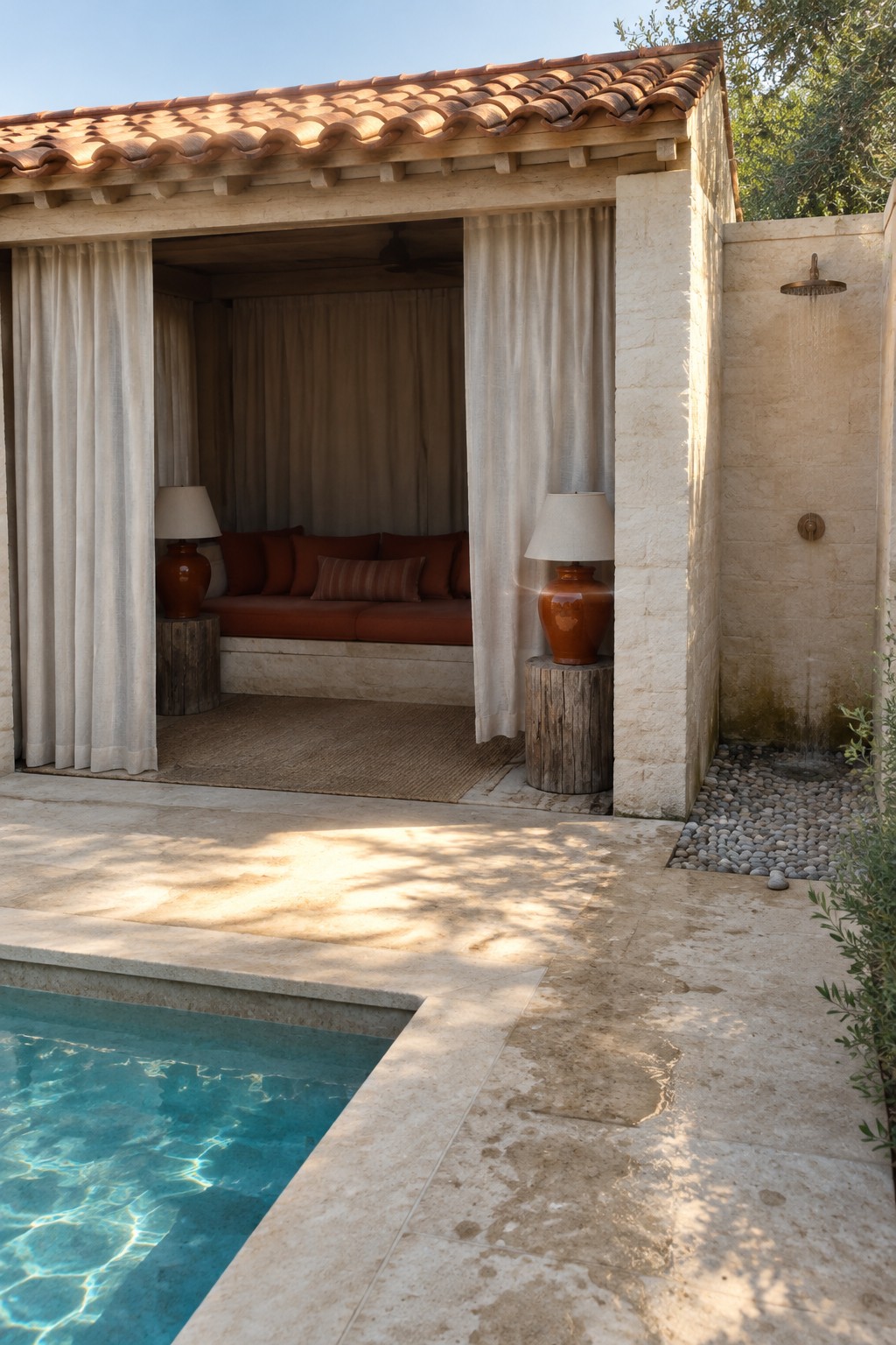 A terracotta-roofed pavilion with a curtained built-in sofa sits beside a stone-paved pool and outdoor shower.