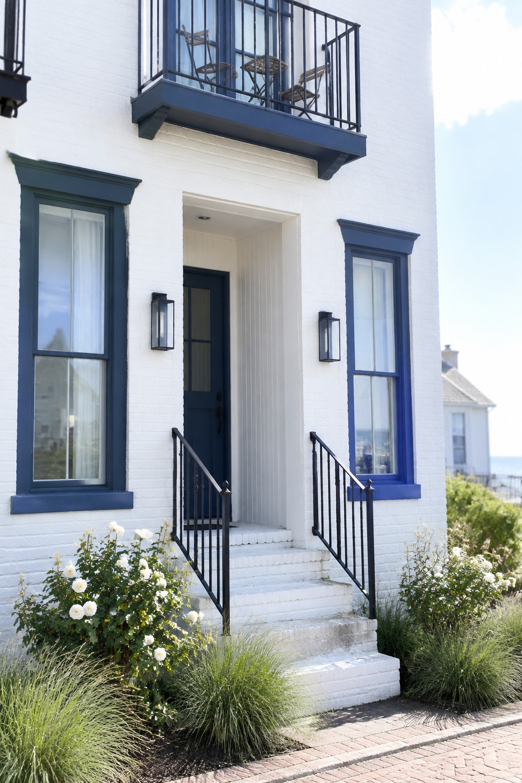White brick coastal house with a recessed dark blue front door, blue-trimmed windows, black railings, and white steps.