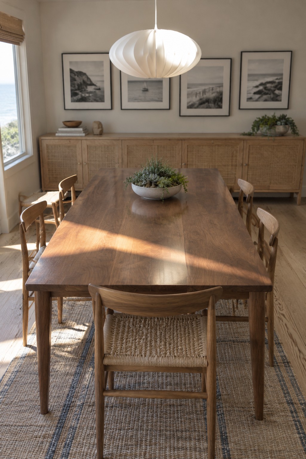 A rectangular wooden dining table with chairs and a woven rug sits beneath a white pendant light in a coastal-style dining room.