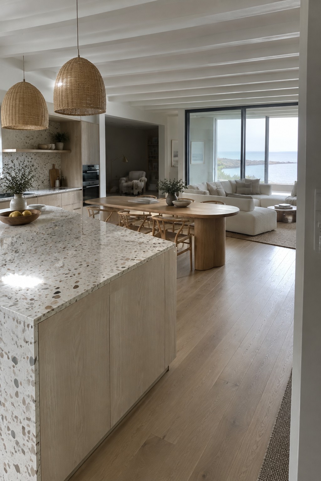 An open-plan coastal interior with a terrazzo kitchen island, long wooden dining table, white sofa, and large windows overlooking the water.